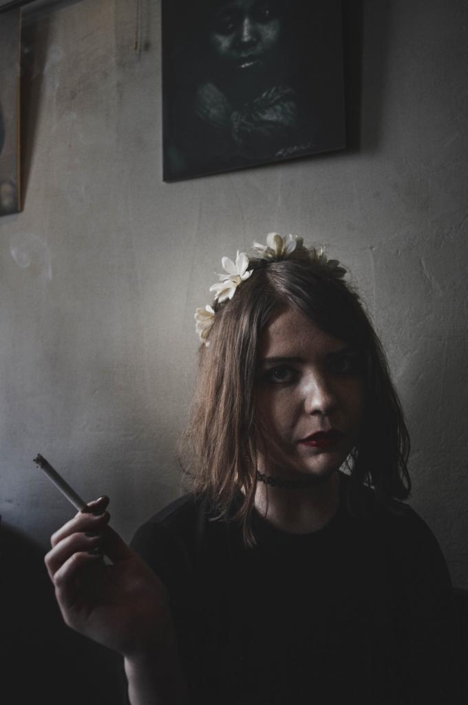 Młoda kobieta z wiankiem z białych kwiatów na głowie, trzymająca zapalonego papierosa, na tle szarej ściany z wiszącymi obrazami.