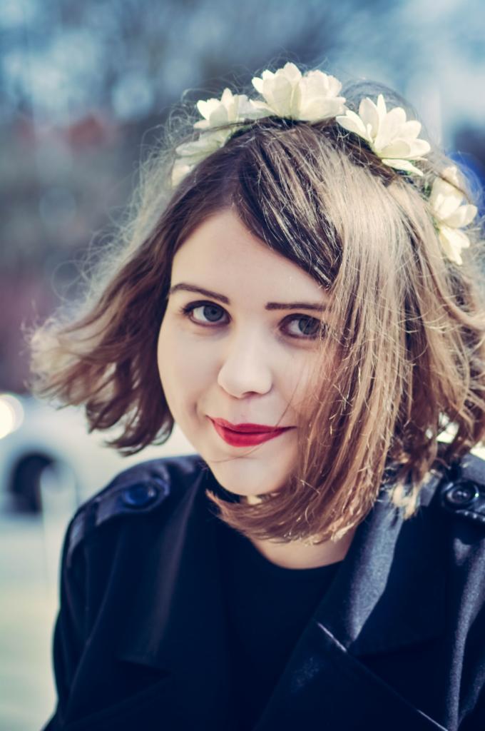 Portret uśmiechniętej kobiety z wiankiem z białych kwiatów na włosach, z wyrazistą czerwoną szminką, w ciemnej bluzce, na rozmytym tle.