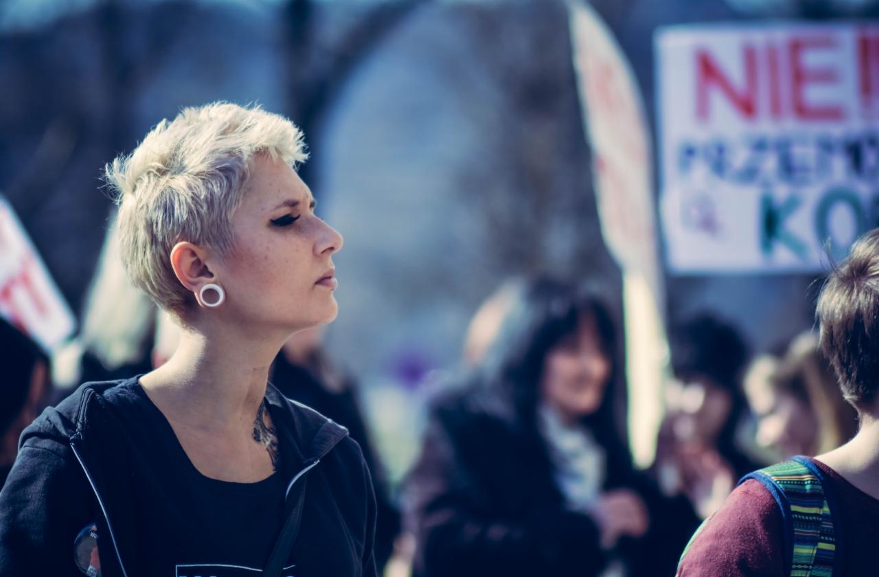 Portret kobiety z krótkimi blond włosami, tunelem w uchu i tatuażem na szyi, spoglądającej w lewo na tle tłumu i transparentów podczas demonstracji.
