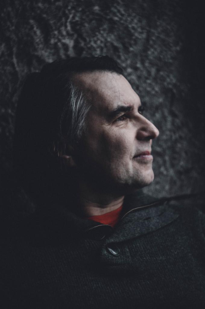 Portret profilowy mężczyzny w szarym swetrze na tle teksturowanej ściany, z widoczną czerwoną koszulką pod spodem, oświetlony bocznym światłem.