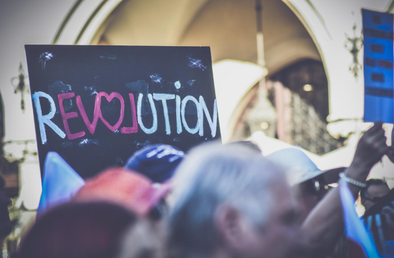 Tłum osób z transparentem 'REVOLUTION' z sercem zamiast litery 'V', napisanym kredą na czarnej tablicy, w tle zamazane architektoniczne detale.