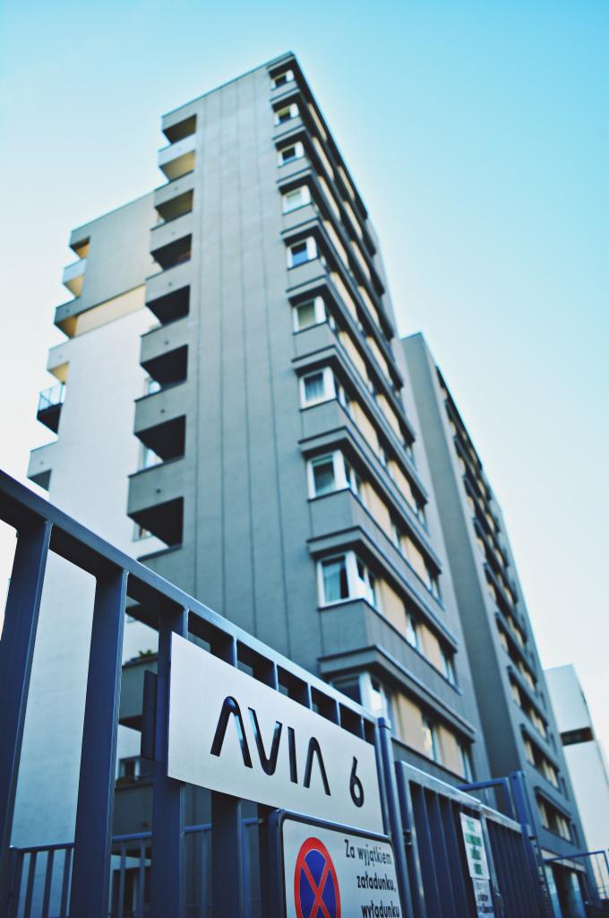 Wysoki, szary budynek mieszkalny z balkonami, widziany z dołu, z tablicą informacyjną 'AVIA 6' na pierwszym planie, umieszczoną na metalowym ogrodzeniu.