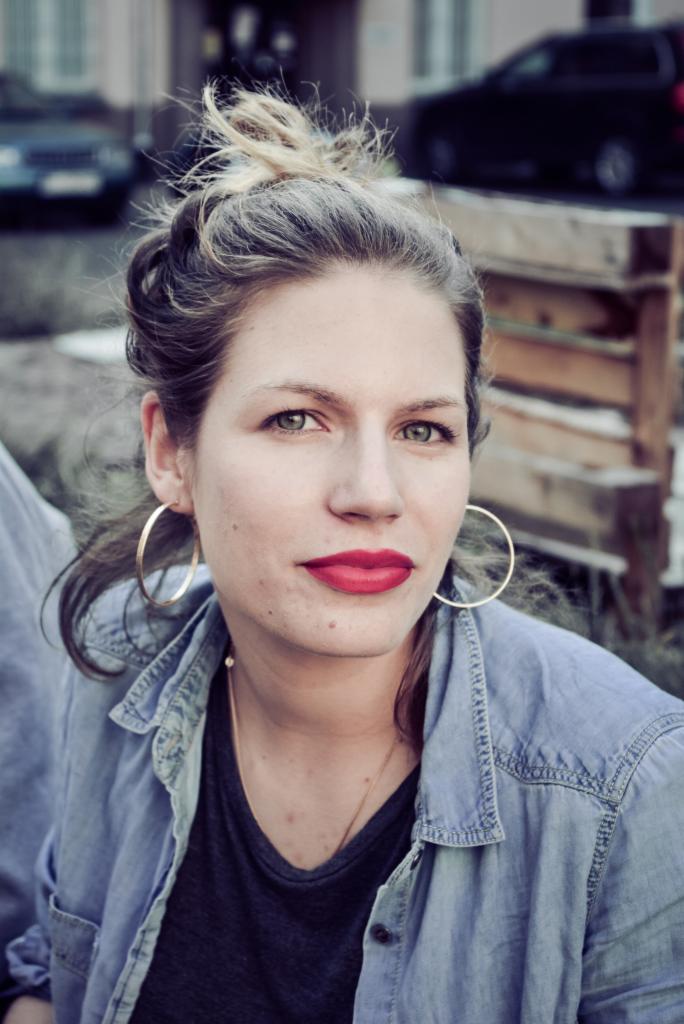 Portret kobiety z włosami upiętymi w luźny kok, noszącej duże złote kolczyki koła i czerwoną szminkę, ubrana w dżinsową koszulę. W tle widoczny drewniany płot i zaparkowane samochody.