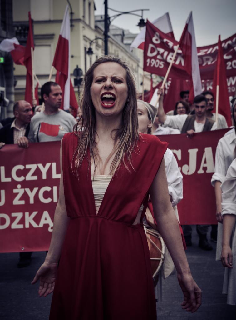 Kobieta w czerwonej sukience krzyczy podczas marszu z czerwonymi flagami i transparentami w tle.