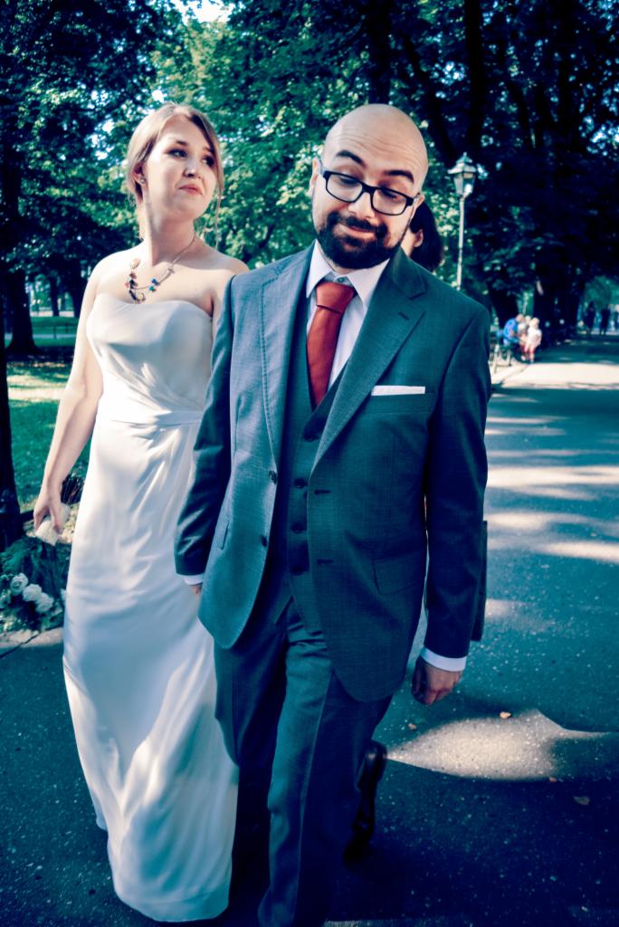 Para młoda spaceruje alejką parkową: panna młoda w białej sukni bez ramiączek i z naszyjnikiem, pan młody w szarym garniturze w kratę, czerwonym krawacie i okularach.