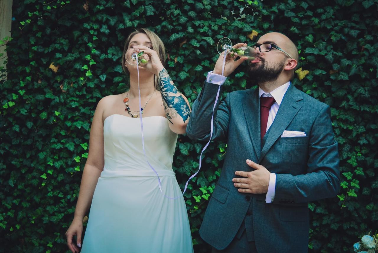 Para młoda wznosi toast szampanem na tle ściany z bluszczu; panna młoda w białej sukni bez ramiączek z tatuażem na ramieniu, pan młody w szarym garniturze i okularach.