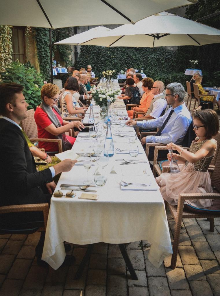 Długi stół z białym obrusem, nakryty dla gości, ustawiony na zewnątrz pod parasolami, z zielonym, porośniętym bluszczem tłem, przy którym siedzą ludzie w eleganckich strojach.