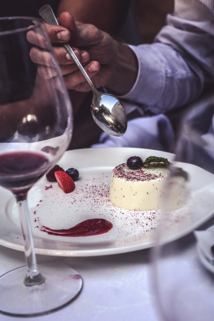 Elegancko podane panna cotta z owocami leśnymi i sosem, odbicie otoczenia w łyżeczce, kieliszek czerwonego wina w tle.