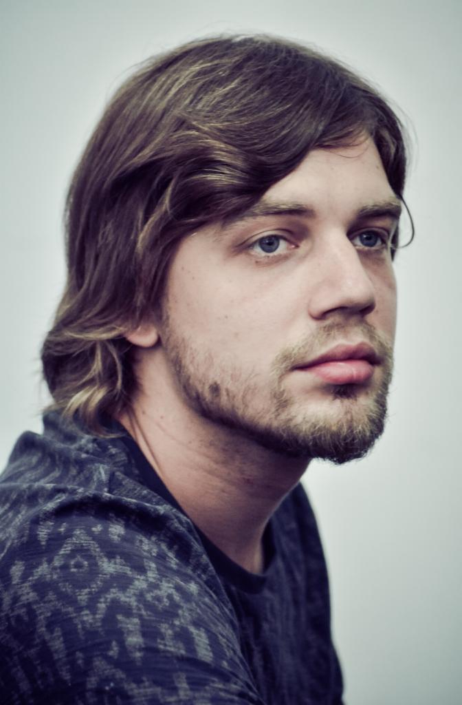 Portret młodego mężczyzny z brązowymi włosami i delikatnym zarostem, skierowanego w lewo, na neutralnym tle.