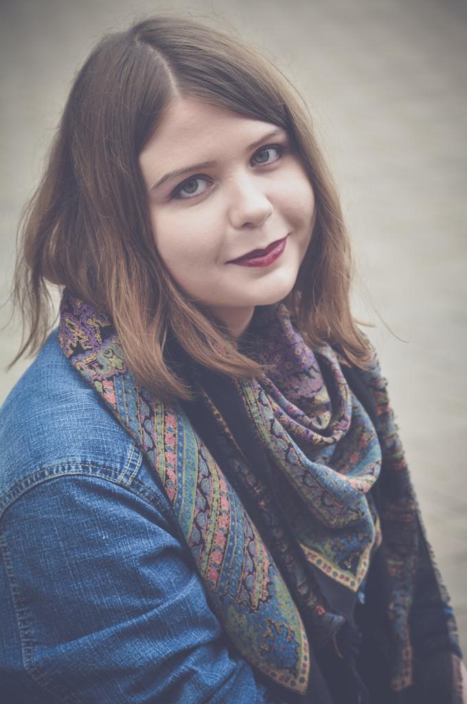 Portret kobiety z ciemną szminką, w dżinsowej kurtce i wzorzystym szalu, z delikatnym uśmiechem i spojrzeniem w górę.