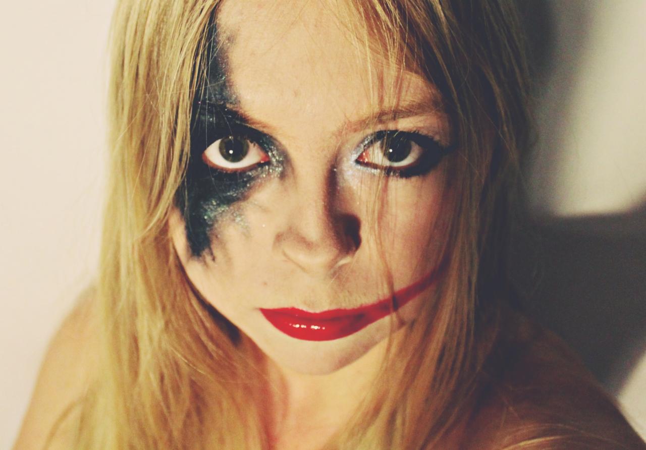 Portret kobiety z awangardowym makijażem: czarne cienie z brokatem na jednym oku i czerwone usta rozciągnięte w szerokim uśmiechu Jokera, jasne włosy opadające na ramiona.
