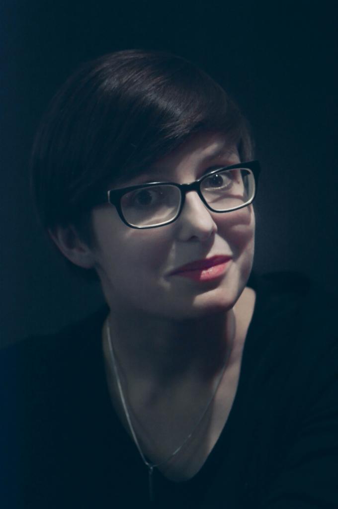 Portret kobiety w okularach z krótkimi ciemnymi włosami i delikatnym makijażem ust, na ciemnym tle.