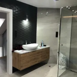 Nowoczesna łazienka z czarną ścianą z trójwymiarową teksturą, białą umywalką nablatową na drewnianej szafce i przeszkloną kabiną prysznicową.