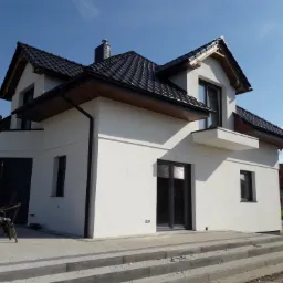 Nowoczesny dom jednorodzinny z czarnym dachem z dachówki ceramicznej i białymi ścianami, widoczny pracownik na rusztowaniu przy balkonie.