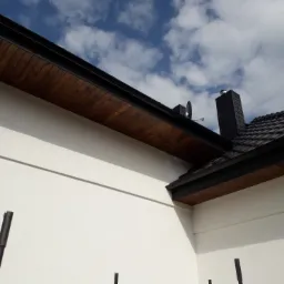 Fragment elewacji domu z nowym, jasnym tynkiem, drewnianym podbitkiem i czarną dachówką oraz kominem, na tle błękitnego nieba z chmurami.