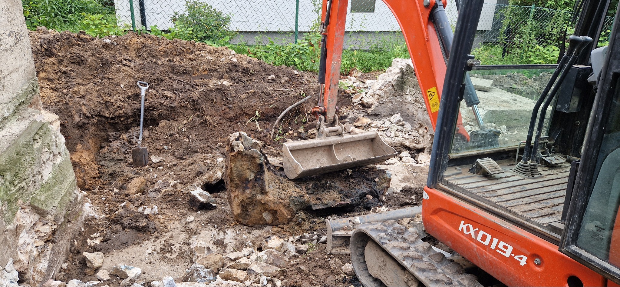 Pomarańczowa mini koparka Kubota KX019-4 w trakcie prac ziemnych, widoczny wykop, fragment starego fundamentu i łopata wbita w ziemię.