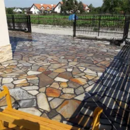 Nieregularny wzór kamiennej nawierzchni na podjeździe, widok z niskiej perspektywy, z elementami drewnianej ławki w rogu kadru i metalowym ogrodzeniem w tle.