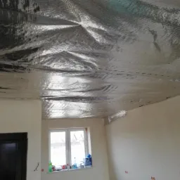 Folia termoizolacyjna z widocznymi szwami na suficie poddasza, surowe ściany w tle, okno z butelkami i pudełkami na parapecie.