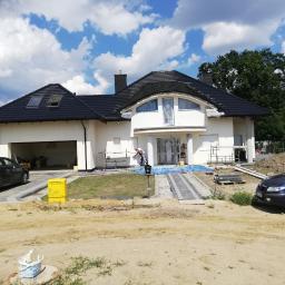 Bud-Tynk - Dwukondygnacyjny dom jednorodzinny z czarnym dachem w trakcie malowania elewacji na biało, widoczne rusztowania i zabezpieczenia okien folią, na pierwszym planie zaparkowane samochody oraz żółta...