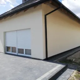 Elewacja budynku z jasnym tynkiem, roletami zewnętrznymi i wykończonym chodnikiem z kostki brukowej.