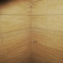 Bud-Tynk - Fragment wykończonej ściany w narożniku łazienki z beżowymi płytkami imitującymi marmur, z dekoracyjnym pasem płytek z ornamentem, widoczne otwory na baterię.