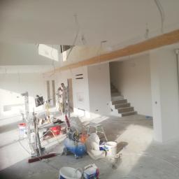 Bud-Tynk - Wnętrze w trakcie malowania natryskowego, widoczne rusztowania, kompresor, wiadra z farbą i malarz w kombinezonie na drabinie, surowy stan wykończenia schodów i belek stropowych.