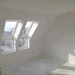 Bud-Tynk - Poddasze po szpachlowaniu i malowaniu na biało, widoczne dwa okna dachowe z widokiem na zewnątrz, surowy beton na podłodze, wiszący kabel elektryczny i otwór na oświetlenie w suficie.