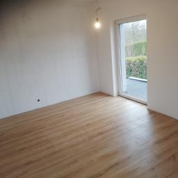 Bud-Tynk - Wykończony pokój z panelami podłogowymi w odcieniach brązu, białe ściany po szpachlowaniu, widoczne kable oświetleniowe oraz okno z widokiem na zieleń na zewnątrz.