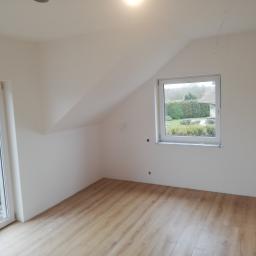 Bud-Tynk - Poddasze po remoncie z oknem i drewnianą podłogą, widoczne ślady po szpachlowaniu na ścianach, przygotowane do dalszych prac wykończeniowych.