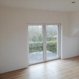 Bud-Tynk - Pusty pokój z jasnymi panelami podłogowymi i dużym oknem balkonowym z widokiem na zieleń ogrodu, świeżo pomalowane ściany na biało, sufit ze skośną ścianą i wystającym kablem.