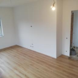 Bud-Tynk - Pokój w trakcie wykańczania: jasne panele podłogowe imitujące drewno, białe ściany z widocznymi punktami elektrycznymi, surowe wykończenie framugi drzwiowej, żarówka na kablu zwisająca z sufitu.