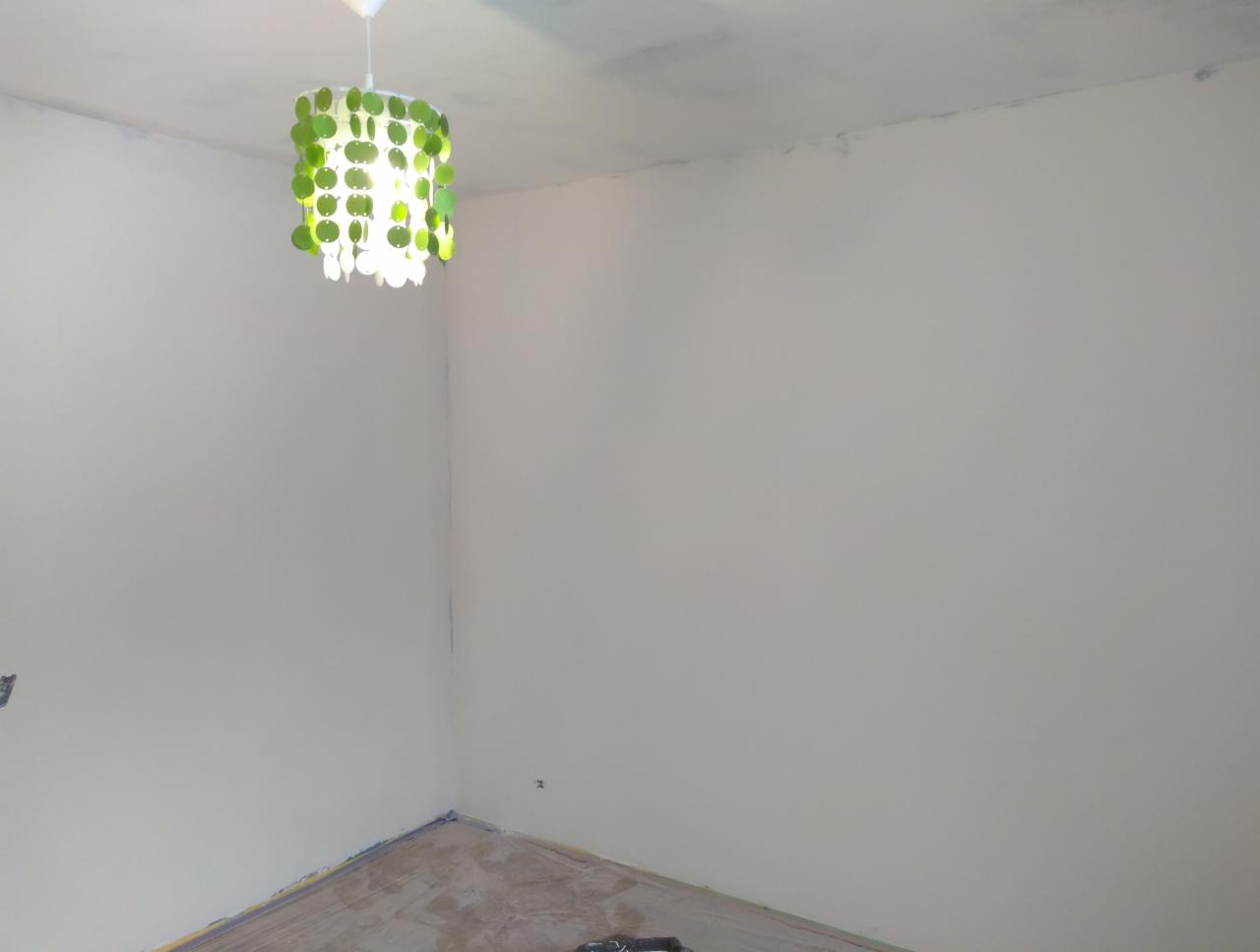 Pokój po szpachlowaniu, z widocznymi śladami po taśmie malarskiej na suficie i podłodze, oraz wiszącą lampą z zielonymi elementami.