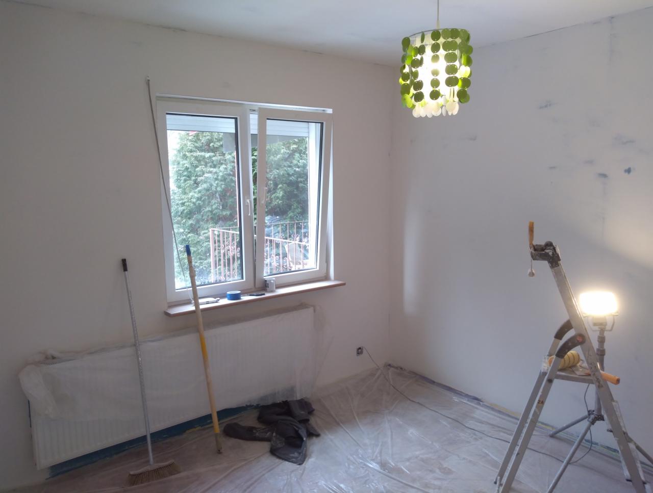 Pokój w trakcie przygotowania do malowania: ściany z widocznymi śladami po szpachlowaniu, zabezpieczona folią podłoga, drabina z lampą halogenową, grzejnik osłonięty folią, okno z widokiem na zieleń.