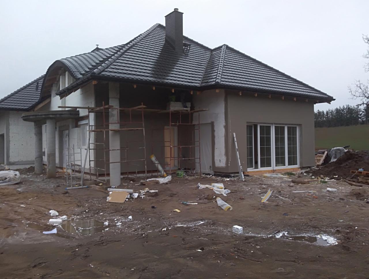 Dom jednorodzinny w trakcie prac wykończeniowych elewacji, widoczne rusztowania, częściowo otynkowana ściana i czarna dachówka na dachu. Na pierwszym planie błotniste podłoże z resztkami materiałów...