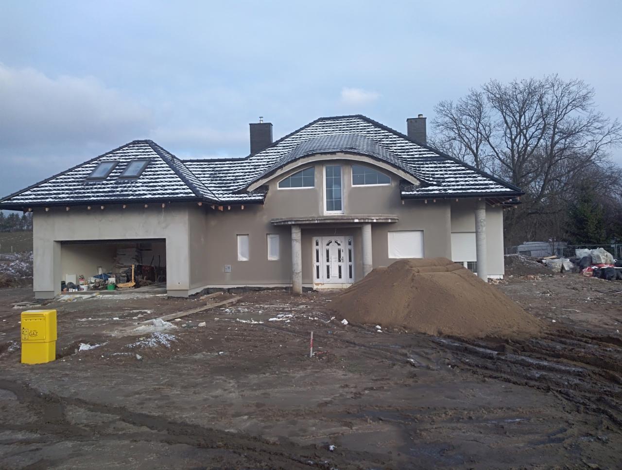 Dom jednorodzinny w trakcie prac wykończeniowych elewacji, z widocznym stosem piasku przed wejściem i otwartym garażem z narzędziami.