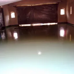 Świeżo wylana, gładka posadzka w pomieszczeniu z oknami, odbijająca światło, zabezpieczona folią na ścianach i drzwiach.