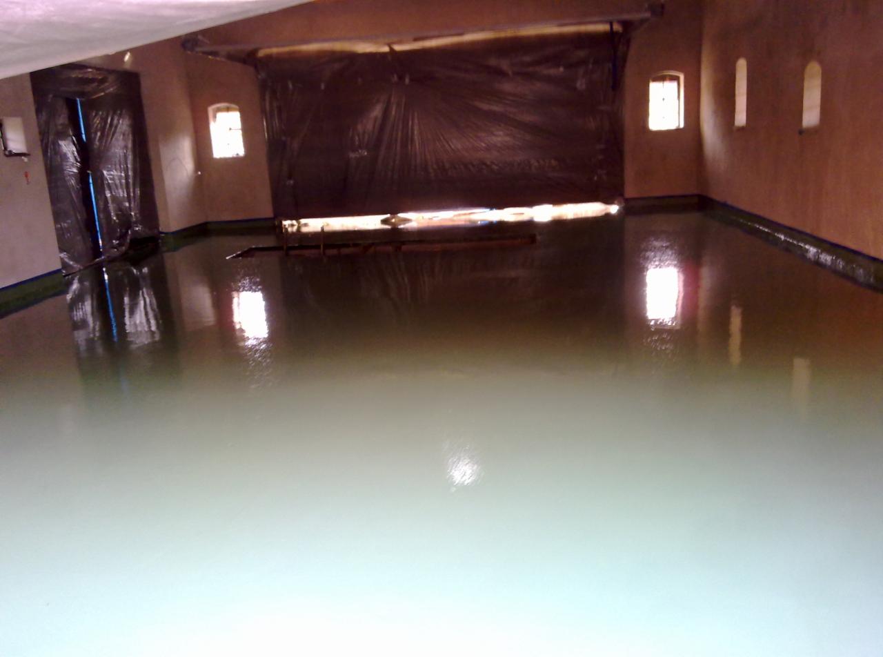 Świeżo wylana, gładka posadzka w pomieszczeniu z oknami, odbijająca światło, zabezpieczona folią na ścianach i drzwiach.