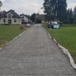 Szeroki, szary podjazd z utwardzonej nawierzchni prowadzący do domu, zabezpieczony po bokach białą folią, z samochodem zaparkowanym obok na trawniku w słoneczny dzień.