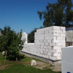 Wznoszenie ścian budynku z białych bloczków betonowych, widoczne elementy konstrukcyjne okna i drzwi, na pierwszym planie paleta styropianu.