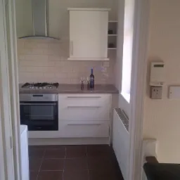 Widok na wykończoną kuchnię z białymi szafkami, brązowymi płytkami podłogowymi, srebrnym okapem i butelką wina z dwoma kieliszkami na blacie, widzianą zza framugi drzwi.