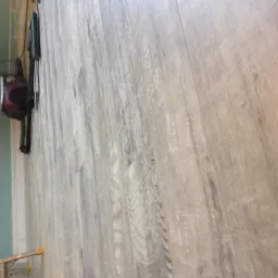 Ułożone panele podłogowe w odcieniach szarości z widocznym fragmentem ściany w kolorze miętowym i gniazdkiem elektrycznym oraz elementem drabiny w tle.