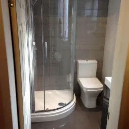 Wykończona łazienka z kabiną prysznicową narożną, toaletą kompaktową i szarą glazurą imitującą drewno, widok zza otwartych drzwi.