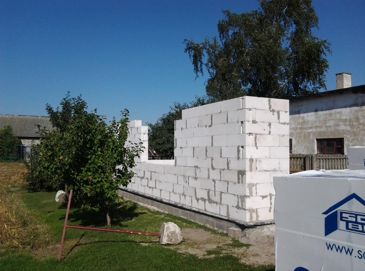 Wznoszenie ścian budynku z białych bloczków betonowych, widoczne elementy konstrukcyjne okna i drzwi, na pierwszym planie paleta styropianu.