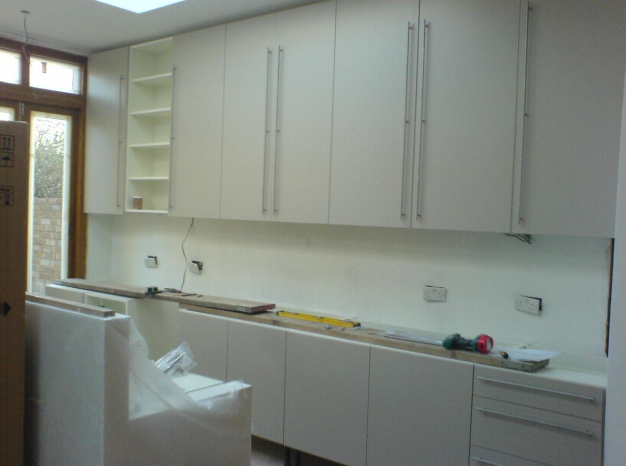 Montaż białych szafek kuchennych z długimi, pionowymi uchwytami, widoczne niezabezpieczone przewody, poziomica, wiertarka akumulatorowa i kartonowe opakowania w pomieszczeniu z oknem dachowym.