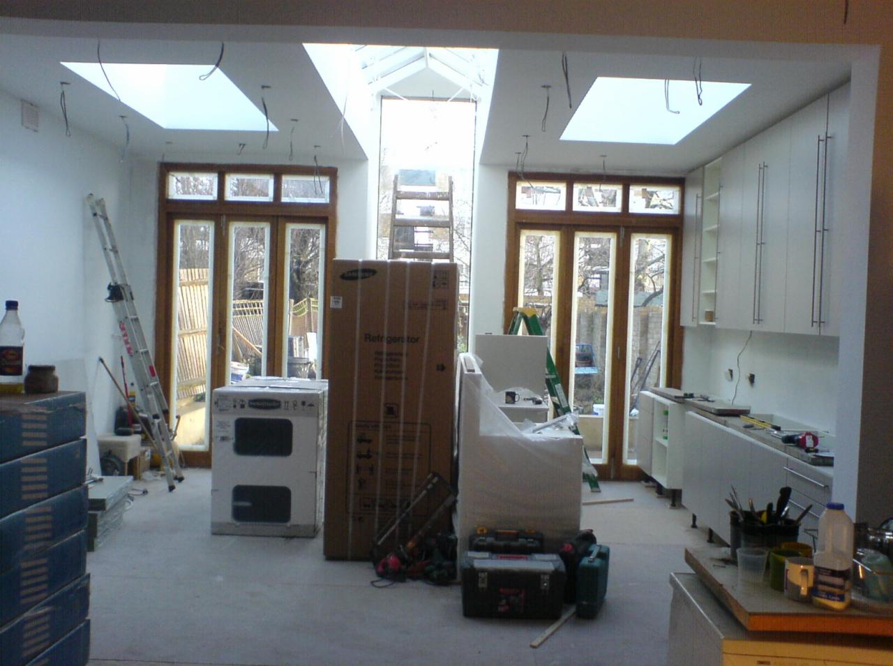 Wnętrze kuchni w trakcie remontu z widocznymi elementami: nowa zabudowa kuchenna, sprzęt AGD w kartonach, drabina, okna balkonowe i świetliki dachowe.