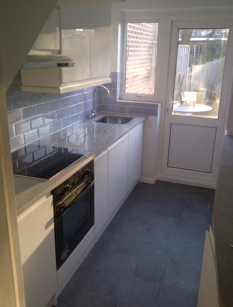 Nowoczesna kuchnia z białymi szafkami, szarymi płytkami metra na ścianie i szarą podłogą, widok na ogród przez drzwi balkonowe.