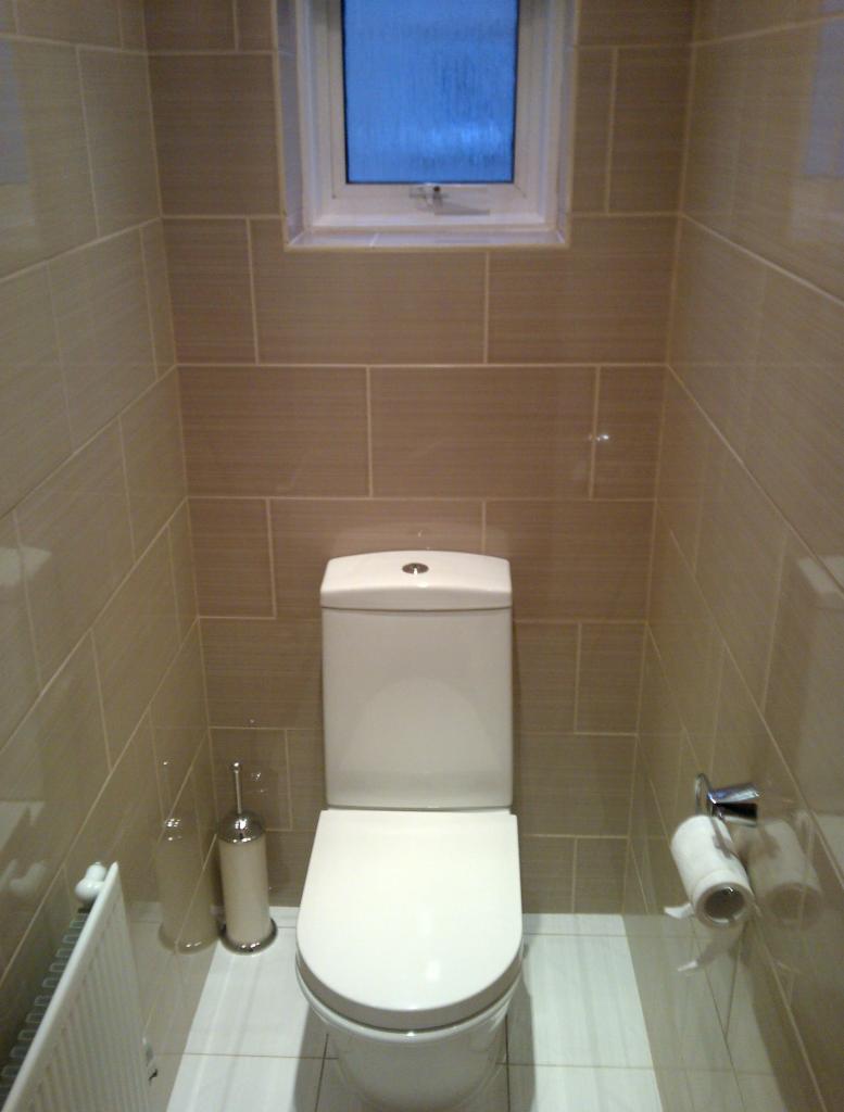 Wykończona toaleta z beżowymi, prostokątnymi płytkami ułożonymi poziomo, małe okno nad spłuczką, biała ceramika sanitarna i grzejnik.