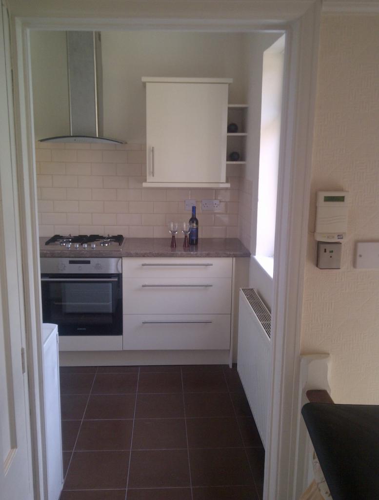 Widok na wykończoną kuchnię z białymi szafkami, brązowymi płytkami podłogowymi, srebrnym okapem i butelką wina z dwoma kieliszkami na blacie, widzianą zza framugi drzwi.