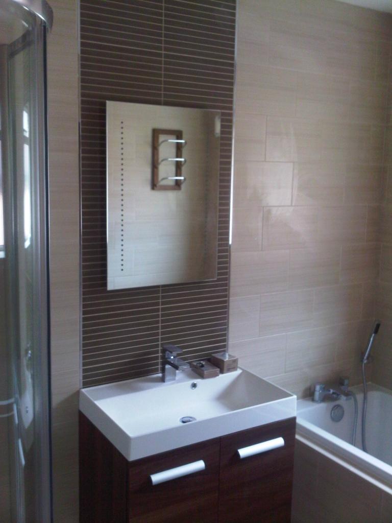 Umywalka z szafką w nowoczesnej łazience, nad nią lustro otoczone ciemnobrązowymi płytkami, obok wanna z baterią.
