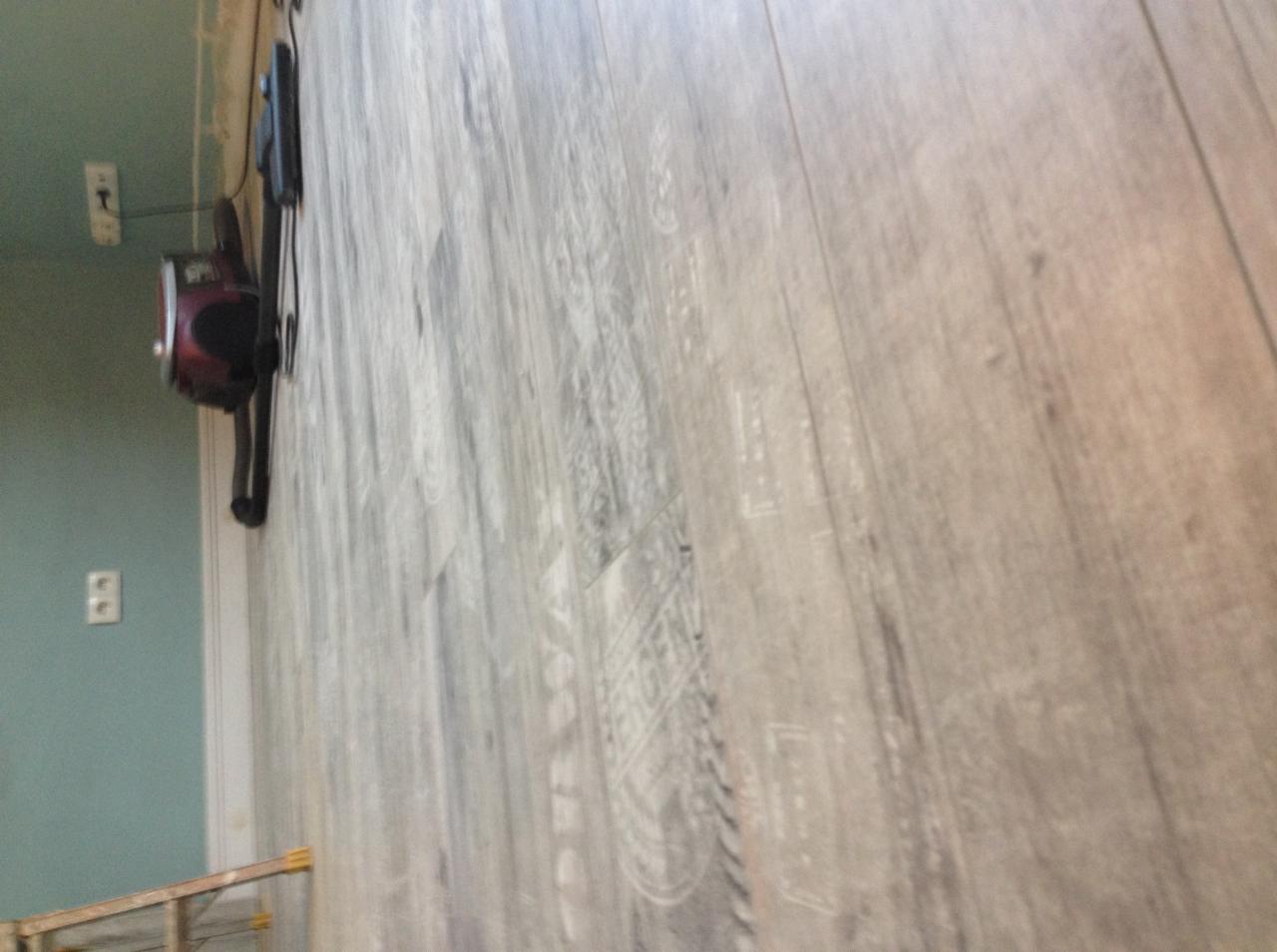 Ułożone panele podłogowe w odcieniach szarości z widocznym fragmentem ściany w kolorze miętowym i gniazdkiem elektrycznym oraz elementem drabiny w tle.